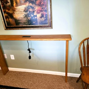 May include: A wooden console table with a light wood finish and a single electrical outlet on the top surface. The table is standing on a carpeted floor in front of a light green wall.