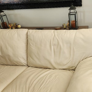 May include: A cream-colored leather sofa with three cushions. The sofa has a smooth, slightly glossy finish. Decorative lanterns and fall foliage are visible on a shelf in the background.