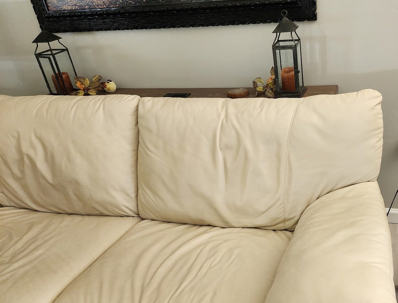 May include: A cream-colored leather sofa with three cushions. The sofa has a smooth, slightly glossy finish. Decorative lanterns and fall foliage are visible on a shelf in the background.