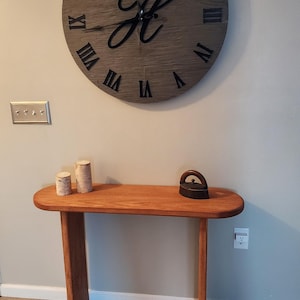 May include: A wooden wall clock with Roman numerals and a cursive letter "H" in the center. The clock is hanging on a white wall above a wooden console table with two candles and a vintage iron.