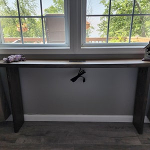May include: A long, wooden console table with a light-colored top and dark gray legs. The table is positioned in front of a window and has a power outlet with a black cord hanging down. A decorative birdhouse sits on the table.