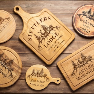 Op de afbeelding: Een verzameling houten snijplanken en onderzetters met gegraveerde ontwerpen. De planken tonen herten, bomen en tekst zoals "Stettlers Lodge" en "Antler Monogram". De items zijn gerangschikt op een houten oppervlak.