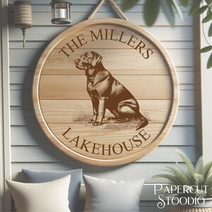 May include: A round wooden sign with the text "The Millers Lakehouse" and a detailed illustration of a dog. The sign is made of light-colored wood and is hanging by a rope.