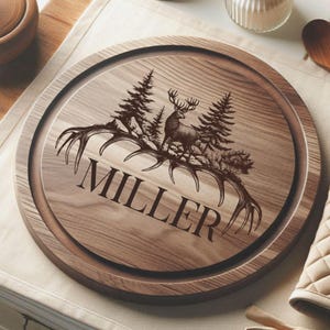 Op de afbeelding: Een ronde houten snijplank met een gegraveerd ontwerp. Het ontwerp toont een hert, dennenbomen en geweien, met de naam "MILLER" eronder. De plank heeft een donkerbruine rand en een lichtere houtnerf in het midden.