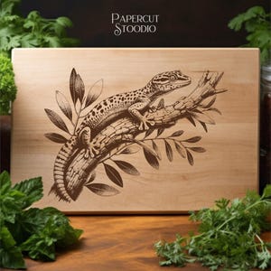 May include: A rectangular wooden cutting board with a detailed brown engraving of a gecko perched on a branch with leaves. The text "PAPERCUT STOODIO" is in the upper right corner. The board has a light wood grain.