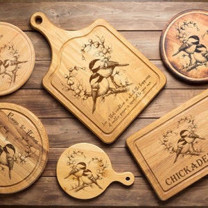 Może przedstawiać: Kolekcja drewnianych desek do krojenia i p&oacute;łmisk&oacute;w do serwowania z grawerowanymi wzorami ptak&oacute;w i kwiat&oacute;w. Niekt&oacute;re elementy mają napis "CHICKADEE LOVE". Przedmioty są ułożone na drewnianej powierzchni.