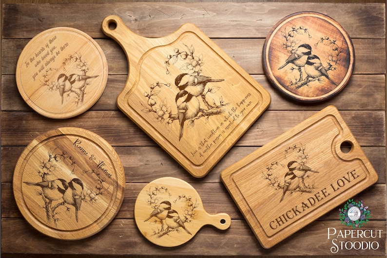 Może przedstawiać: Kolekcja drewnianych desek do krojenia i p&oacute;łmisk&oacute;w do serwowania z grawerowanymi wzorami ptak&oacute;w i kwiat&oacute;w. Niekt&oacute;re elementy mają napis "CHICKADEE LOVE". Przedmioty są ułożone na drewnianej powierzchni.