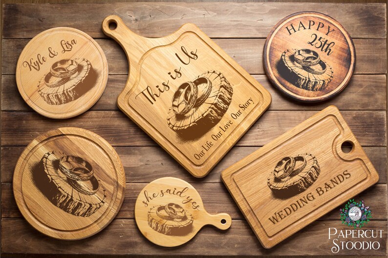 May include: Set of five wooden cutting boards with engraved rings and text. The boards are in various shapes and sizes, including round, square, and rectangular. The text on the boards includes "Kyle & Lisa", "This is Us", "Our Life Our Love Our Story", "She said yes", "Happy 25th", and "Wedding Bands".