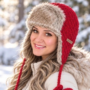 May include: A bright red trapper hat featuring a textured knit pattern and thick, light brown faux fur lining on the forehead and ear flaps. The hat includes long, braided red ties hanging from the sides and a small rectangular brown tag.
