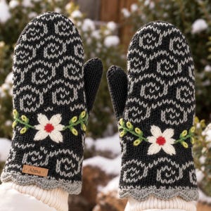 May include: A pair of black and grey knit mittens featuring a repeating swirl pattern, a white flower with a red centre, and green leaves. The mittens have a scalloped grey cuff and a small rectangular brown leather tag with the text Alma.