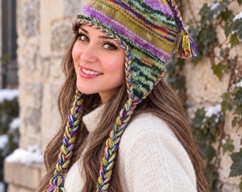 May include: A hand-knit beanie with earflaps and long, thick braided tassels in a variegated yarn featuring shades of purple, green, yellow, and blue. The hat includes a small rectangular brown leather tag with the text ALMA embossed on it.
