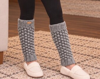 May include: A person wearing a cream-colored chunky knit sweater, black leggings, and grey textured leg warmers with a bobble pattern. The leg warmers feature a small rectangular brown tag at the top. The outfit is completed with white slip-on shoes.