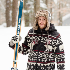 May include: A man wearing a black and white patterned sweater with a fur-trimmed hat holds a pair of vintage wooden skis with blue and white bindings. The skis are labeled "Splitkein".