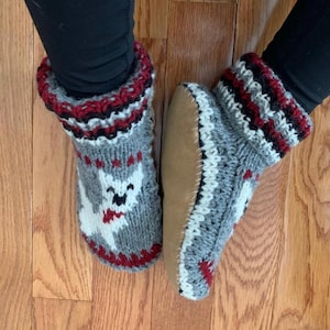 May include: A pair of grey and white knitted slipper socks with a red and black trim. The socks feature a white cartoon animal design.