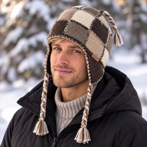 May include: A knit earflap hat featuring a brown, beige, and cream checkered pattern. The hat includes braided tassels hanging from the earflaps and a small braided tassel at the crown. A small rectangular brown tag is visible on the side of the hat.