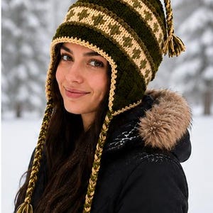 May include: A dark olive green crochet hat featuring a cream-colored geometric pattern, ear flaps, and long braided tassels with matching cream and green fringe at the ends. The hat has a textured stitch and a cream-colored border along the edges.
