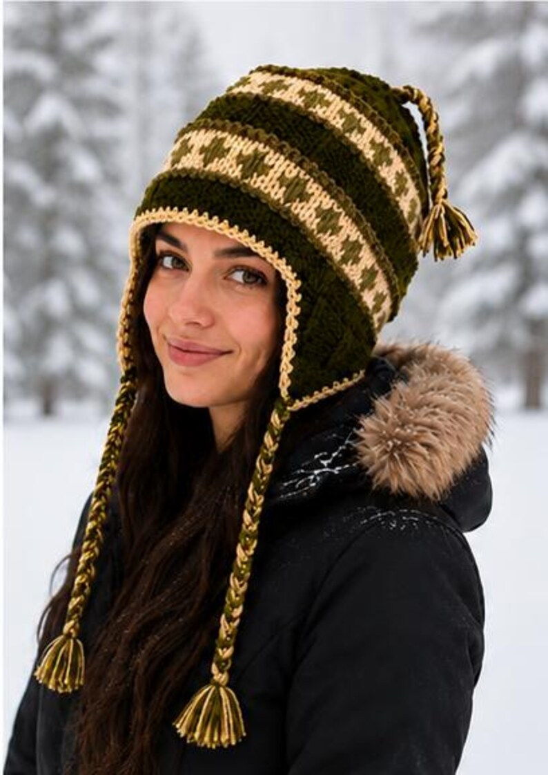 May include: A dark olive green crochet hat featuring a cream-colored geometric pattern, ear flaps, and long braided tassels with matching cream and green fringe at the ends. The hat has a textured stitch and a cream-colored border along the edges.