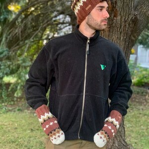 Pu&ograve; includere: Una persona indossa un berretto di lana a maglia con motivo marrone e bianco, una giacca in pile nera con un logo verde e guanti di lana a maglia con motivo marrone e bianco. La persona &egrave; in piedi davanti a un albero.