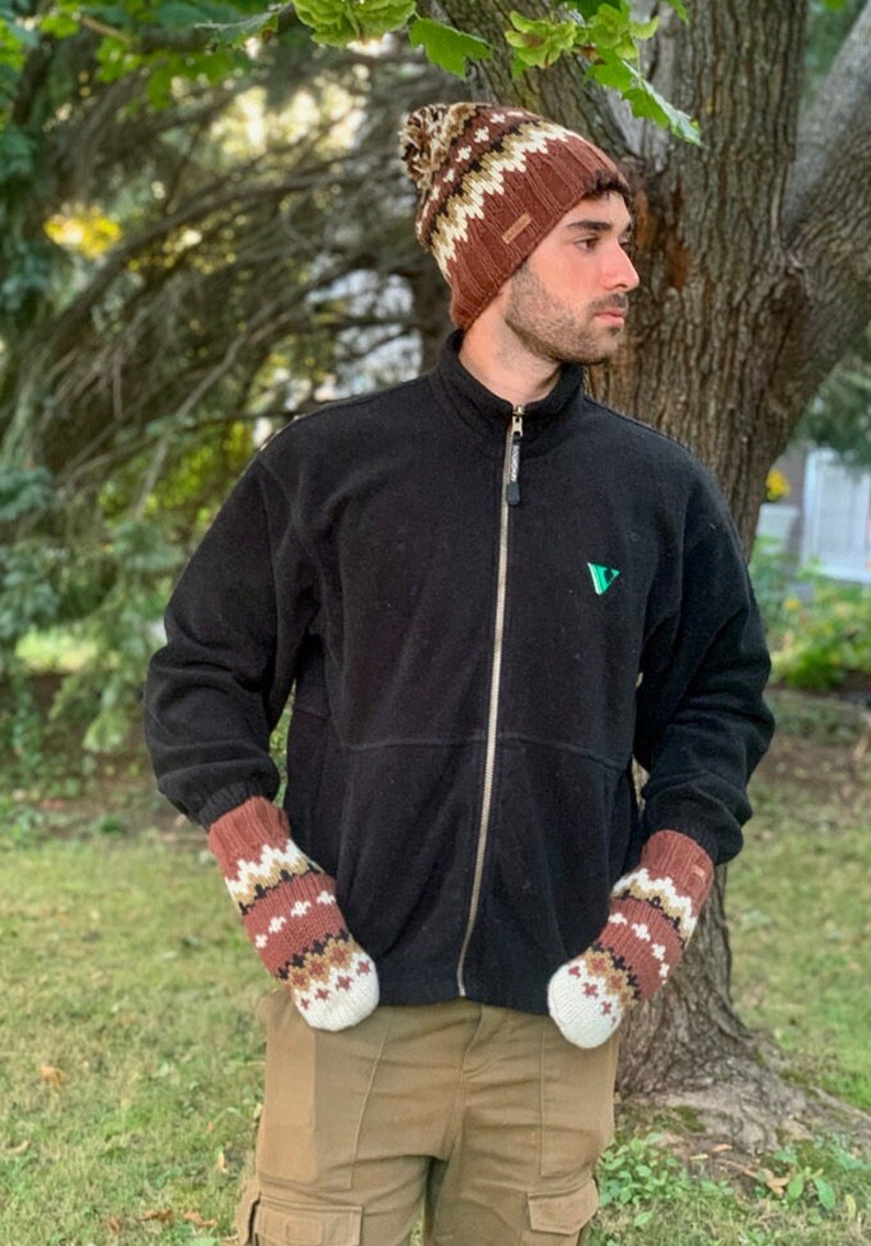Pu&ograve; includere: Una persona indossa un berretto di lana a maglia con motivo marrone e bianco, una giacca in pile nera con un logo verde e guanti di lana a maglia con motivo marrone e bianco. La persona &egrave; in piedi davanti a un albero.