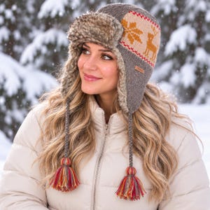 May include: A gray knit trapper hat featuring a beige panel with a repeating orange deer and snowflake pattern. The hat includes a faux fur-lined ear flap, braided side ties, and multicolored yarn tassels at the ends of each braided cord.