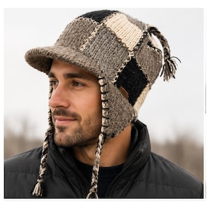 May include: A knit hat featuring a patchwork design in shades of black, gray, and cream. The hat includes a stiff brim, ear flaps, and braided tassels hanging from the sides and top. The panels are joined with visible light-colored stitching.