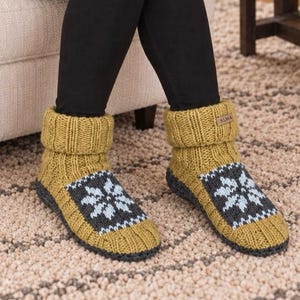 May include: A pair of mustard yellow knit slipper socks featuring a ribbed cuff and a dark grey panel on the top of the foot with a light blue snowflake pattern. A small rectangular brown tag with the text ALMA is attached to the cuff.