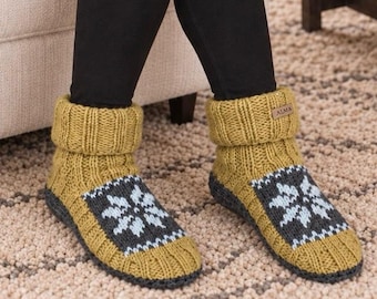 May include: A pair of mustard yellow knit slipper socks featuring a ribbed cuff and a dark gray panel on the top of the foot with a light blue snowflake pattern. A small rectangular brown tag with the text ALMA is attached to the cuff.