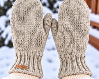 May include: A pair of beige knit mittens featuring a ribbed cuff and a small brown rectangular leather tag with the word ALMA embossed on the left mitten. The mittens are constructed with a thick, textured yarn in a classic stockinette stitch.