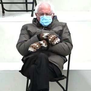 Pu&ograve; includere: Un uomo con una maschera facciale blu e guanti di lana marroni &egrave; seduto su una sedia pieghevole nera. Indossa una giacca grigia e pantaloni neri.