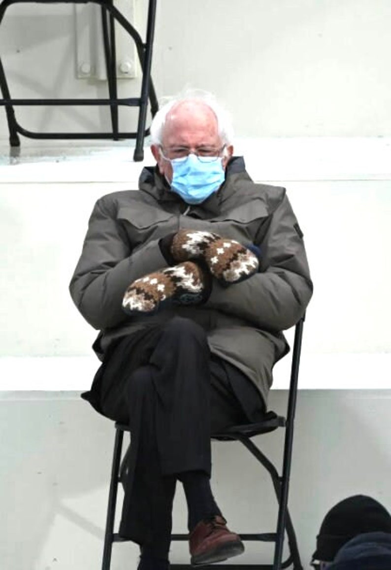 Pu&ograve; includere: Un uomo con una maschera facciale blu e guanti di lana marroni &egrave; seduto su una sedia pieghevole nera. Indossa una giacca grigia e pantaloni neri.