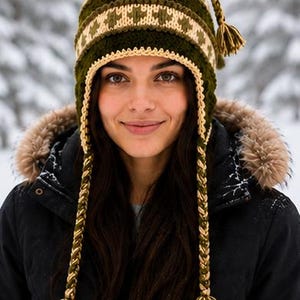 May include: A dark olive green and cream knit hat featuring a geometric pattern, a ribbed texture, and two long braided earflaps that end in thick tassels. The hat includes a matching braided cord with a tassel at the top center.