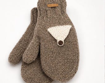 Könnte beinhalten: Ein Paar braune Strickfäustlinge mit geripptem Bündchen und strukturierter Oberfläche. Jeder Fäustling hat einen dreieckigen cremefarbenen Aufnäher auf der Rückseite mit einem kleinen braunen Knopf an der Spitze. Ein beiges Lederetikett trägt den Text ALMA.