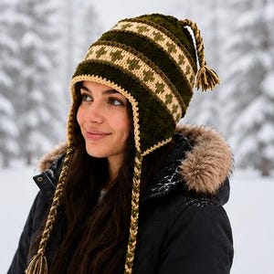 May include: A dark olive green knit hat featuring a cream-colored geometric pattern, a textured stitch design, and long braided earflaps ending in tassels. The hat includes a matching braided top detail with a tassel and a cream-colored trim.