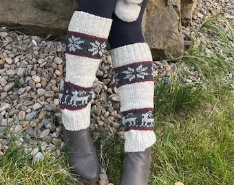 May include: A pair of cream-coloured knit leg warmers featuring a dark grey band with white snowflake patterns and a second dark grey band with white reindeer motifs, accented by thin maroon stripes, worn over black leggings and grey boots.