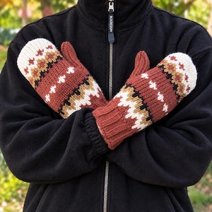 May include: A pair of knit mittens featuring a geometric pattern in shades of rust orange, cream, black, and tan. The mittens have a ribbed cuff and are worn over a black fleece zip-up jacket with a visible zipper pull labelled with the word BOLDIARY.