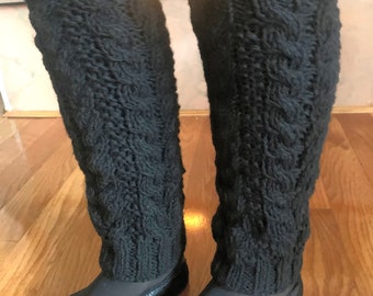 May include: Black knitted leg warmers with a cable knit pattern. The leg warmers are worn over black tights and black shoes.