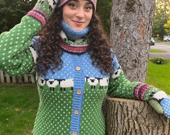 May include: A knitted set consisting of a button-up cardigan, a beanie, a cowl, and mittens. The garments feature a repeating pattern of black and white sheep on a green field under a blue sky, with pink and dark brown accents at the borders.
