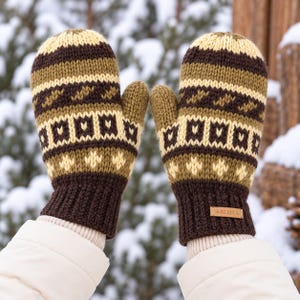 May include: A pair of thick, knit mittens featuring a horizontal pattern of dark brown, olive green, and cream stripes with geometric shapes. The cuffs are ribbed dark brown knit with a small rectangular leather tag labelled ALMA in the centre.