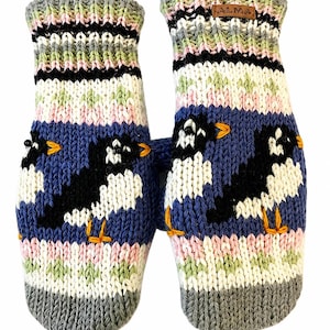 May include: A pair of knitted mittens featuring a blue background with black and white bird motifs, orange beaks and feet, and horizontal stripes in green, pink, white, and black. A small brown leather tag with the text ALMA is attached to one cuff.