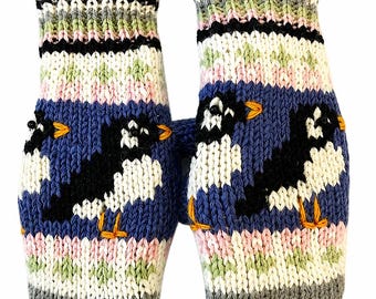 May include: A pair of knit mittens featuring a blue background with black and white bird motifs, orange beaks and feet, and horizontal stripes in green, pink, white, and black. A small brown leather tag with the text ALMA is attached to one cuff.