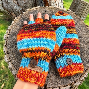 May include: A pair of fingerless convertible mittens featuring a chunky knit pattern with horizontal stripes in vibrant shades of orange, red, brown, and bright blue. Each mitten includes a small dark brown wooden button and a rectangular leather tag.
