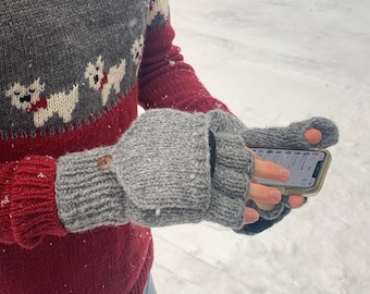 May include: A pair of gray fingerless gloves with a brown button on the cuff. The gloves are made of a soft, knitted fabric and have a fingerless design. The person is wearing the gloves and using a smartphone.