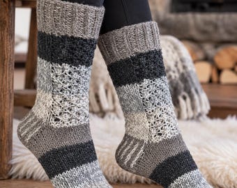 May include: A pair of mid-calf knit socks featuring a color-blocked pattern in shades of charcoal, light gray, and white. The socks display a combination of ribbed cuffs, solid knit sections, and an intricate openwork lace stitch pattern throughout.
