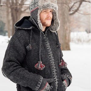 May include: A dark charcoal grey knit hooded jacket featuring a front zip, white contrast stitching, and two hanging tassels. The jacket is paired with a matching grey knit hat with a faux fur trim and a red and white patterned design.
