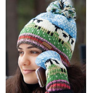 May include: A knit beanie with a pom-pom and matching mittens featuring a landscape pattern of green hills, black sheep, and a blue sky. The beanie has a brown rectangular tag with the text ALMA. The design includes bands of red and purple.