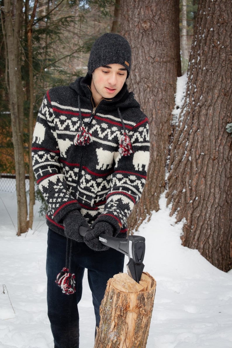 May include: A person wearing a black and white patterned sweater and a black beanie hat is chopping wood with a black axe. The person is standing in a snowy forest setting.