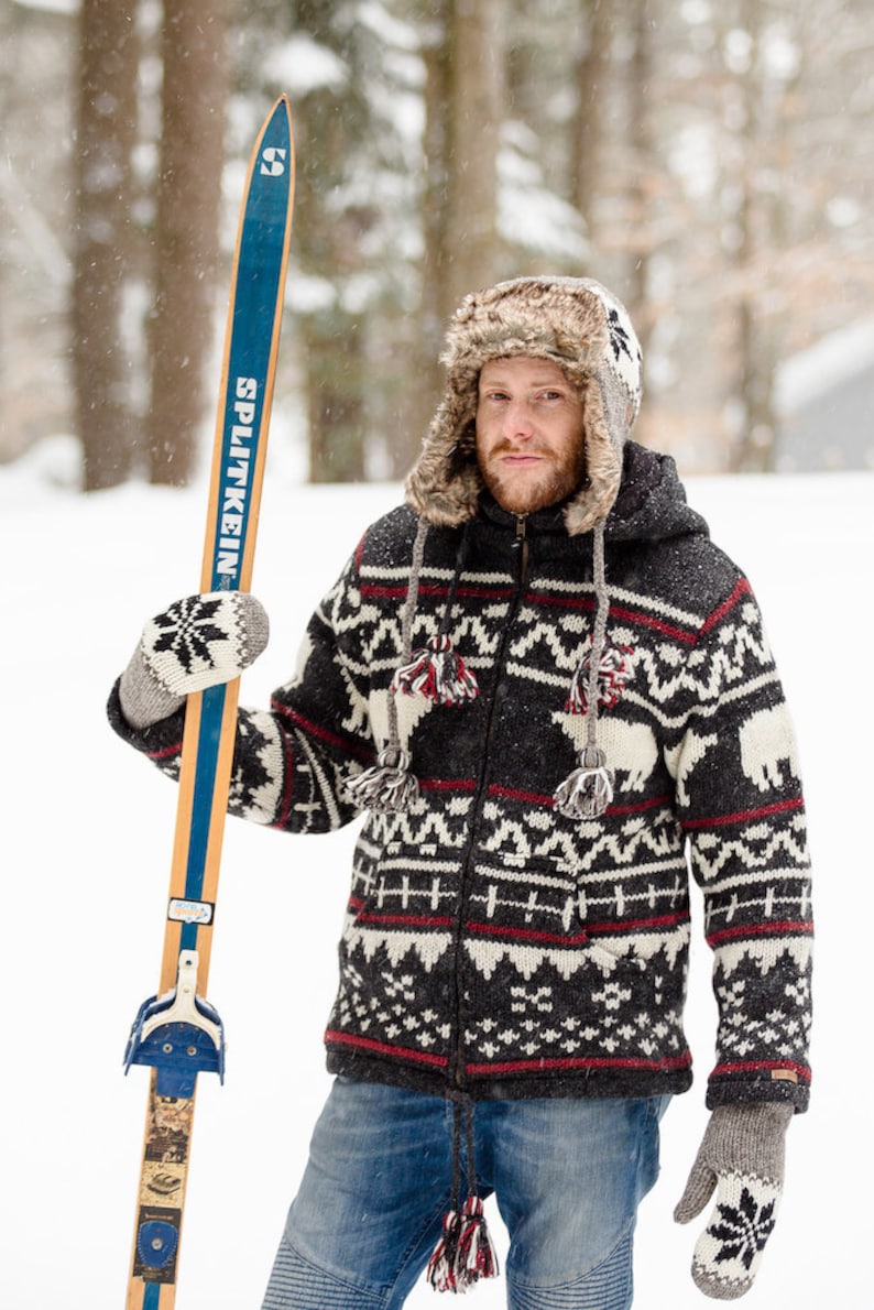 May include: A man wearing a black and white patterned sweater with a fur-trimmed hat holds a pair of vintage wooden skis with blue and white bindings. The skis are labeled "Splitkein".
