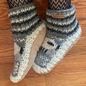 May include: A pair of black, white and grey knitted slipper socks with a brown leather label that says "ALMA".