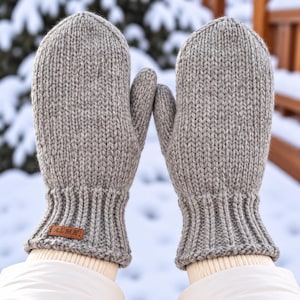 May include: A pair of light grey knit mittens featuring a ribbed cuff and a small rectangular brown leather tag with the embossed text ALMA. The mittens are constructed with a thick, chunky yarn texture and have a rounded thumb and finger section.