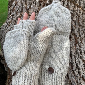 Puede incluir: Un par de mitones convertibles de punto grueso en color gris claro, con diseño sin dedos y una solapa plegable. Cada mitón está decorado con un botón circular de color marrón oscuro en el dorso de la mano y una pequeña etiqueta rectangular.